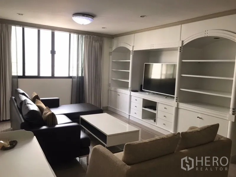 Condominium for rent in Watthana, Bangkok - Bright living room with large windows, a white built‑in media wall with shelving and TV, black leather sectional and coffee table.
