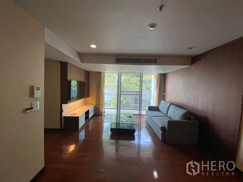 คอนโด ให้เช่าในWatthana, Bangkok - ห้องนั่งเล่นกว้างพร้อมโซฟา ผนังทีวี พื้นไม้ และประตูกระจกสไลด์ออกสู่ระเบียงวิวต้นไม้