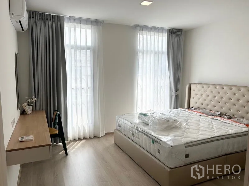 ทาวน์เฮาส์ ให้เช่า - ห้องนอนใหญ่โปร่งโล่ง มีหน้าต่างสูงสองบาน โต๊ะทำงาน และเตียงหุ้มบุ.