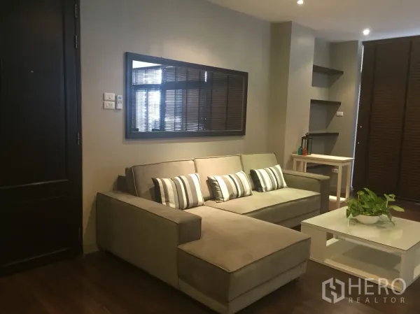 อพาร์ทเมนท์ ให้เช่าในKhlong Toei, Bangkok - ห้องนั่งเล่นโทนอบอุ่น โซฟาเข้ามุม หมอนลายทาง กระจกเงาบนผนัง และโต๊ะกลางสีขาว