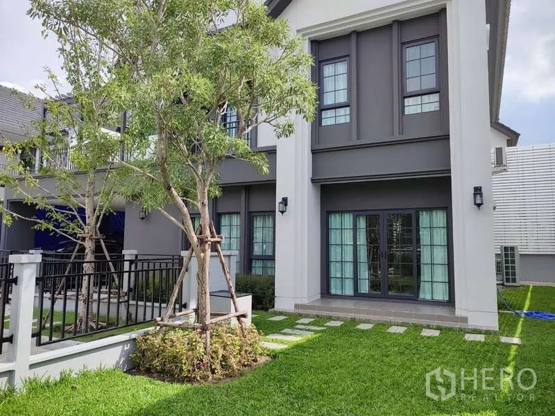 Detached House for rent in Bang Phli, Samut Prakan - Side exterior showing lawn, French doors, trees and metal fence.