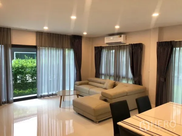 Detached House for rent in Don Mueang, Bangkok - Open-plan living room with L-shaped leather sofa, sheer curtains and air conditioner.