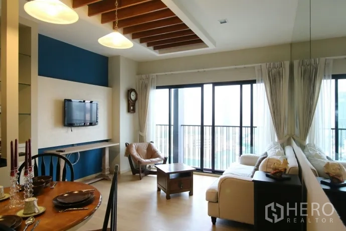 Condominium for rent or sale in Watthana, Bangkok - Open-plan living room with large sliding glass doors, sofa, TV and city-view balcony railing.