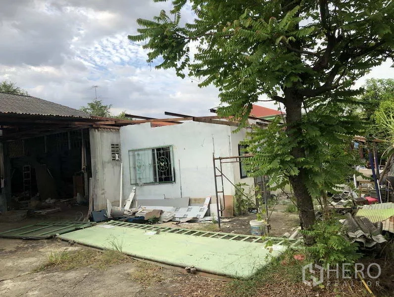 คลังสินค้า ให้เช่าในSai Mai, Bangkok - มุมด้านข้างโกดังพร้อมอาคารเล็ก ต้นไม้ร่มรื่น และวัสดุก่อสร้างกระจายอยู่