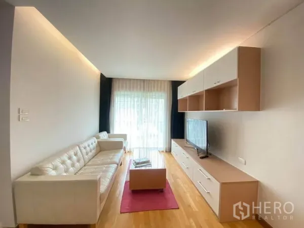Condominium for rent or sale in Phra Khanong, Bangkok - Light-filled living room with white sofa, TV wall unit and sliding glass door to the balcony with sheer curtains.