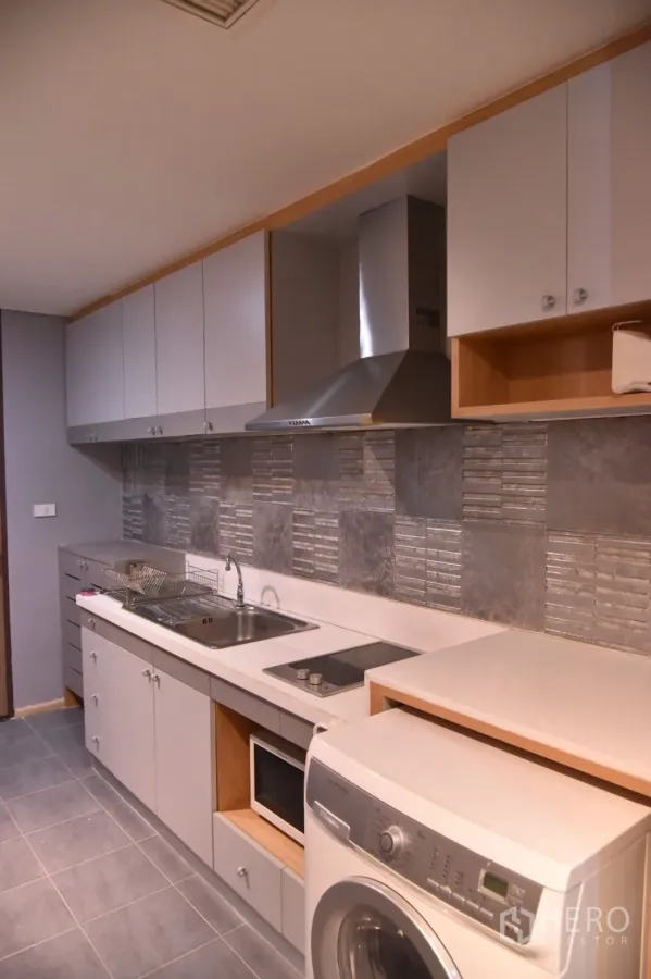 Condominium for rent or sale in Khlong Toei, Bangkok - Modern galley kitchen with grey cabinets, tiled backsplash, cooktop, hood, microwave and front-load washer.