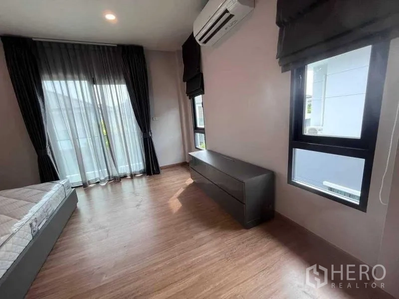 Detached House for rent in Don Mueang, Bangkok - Sunlit bedroom with air conditioner, balcony door and low cabinet by windows.