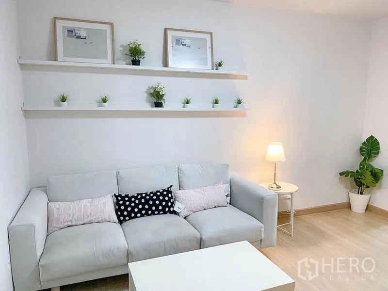 คอนโด ขายในKhlong Toei, Bangkok - ห้องนั่งเล่นพร้อมโซฟานุ่ม ชั้นวางของสีขาวตกแต่งต้นไม้ และโคมไฟโต๊ะข้าง