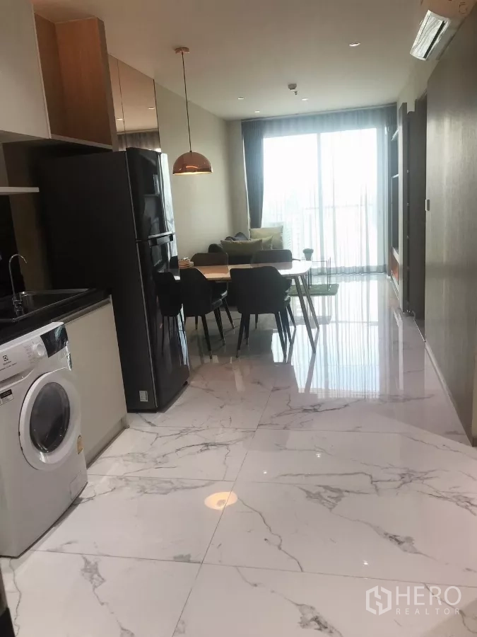 Condominium for rent in Watthana, Bangkok - Hallway with marble‑look floors, fitted kitchen, fridge and dining set leading to a bright balcony.