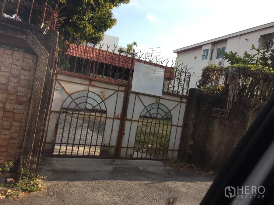 Detached House for sale in Watthana, Bangkok - Gated entrance with rusted iron fence and concrete wall of a detached house in Watthana, Bangkok, showing driveway and mature trees.