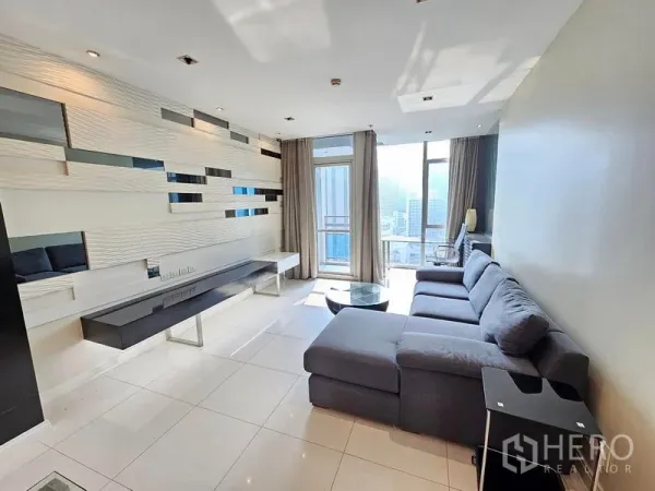 Condominium for rent or sale in Pathum Wan, Bangkok - Bright living room with L-shaped sofa, feature wall and floor-to-ceiling window opening to a balcony with city views.