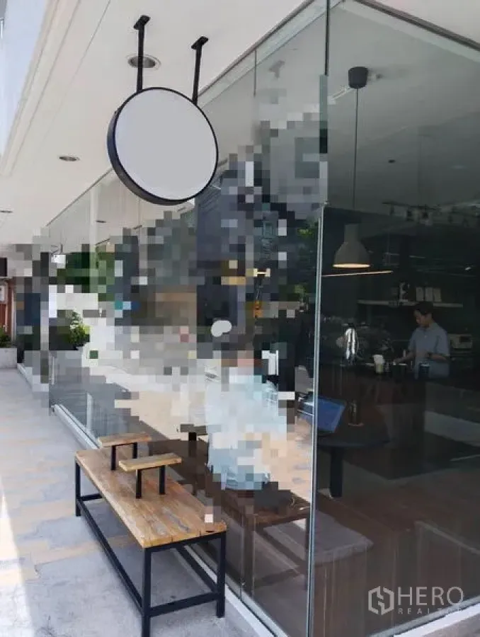 零售空间 出租在Watthana, Bangkok - 临街玻璃店面，圆形悬挂招牌与木质长凳，行人通道可见。