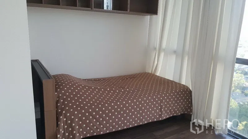 คอนโด ให้เช่าในBangkok - ห้องนอนขนาดกะทัดรัด มีเตียงเดี่ยว ผ้าม่านโปร่ง และหน้าต่างเต็มบานมองวิวเมือง