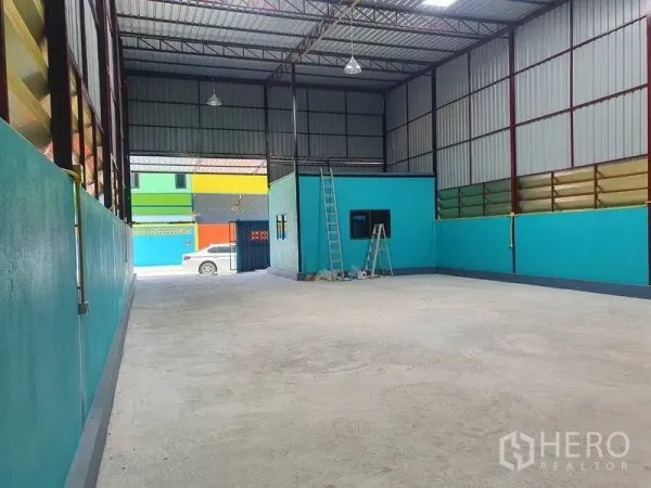 Warehouse for rent in Nonthaburi - High-ceiling warehouse interior with steel roof, skylights, teal walls, wide concrete floor and an internal office block.