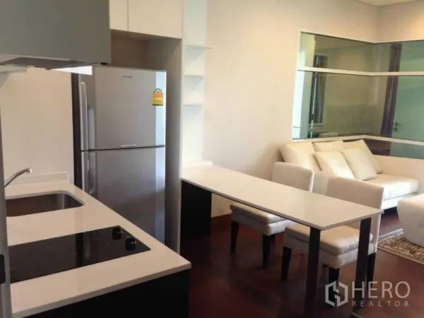 Condominium for rent or sale in Watthana, Bangkok - Open-plan Thonglor condo with compact kitchen, induction cooktop, stainless fridge and dining counter facing a bright living area with white sofa and glass partition.