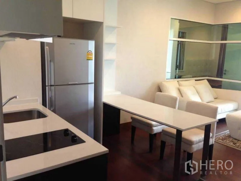 Condominium for rent or sale in Watthana, Bangkok - Open-plan Thonglor condo with compact kitchen, induction cooktop, stainless fridge and dining counter facing a bright living area with white sofa and glass partition.