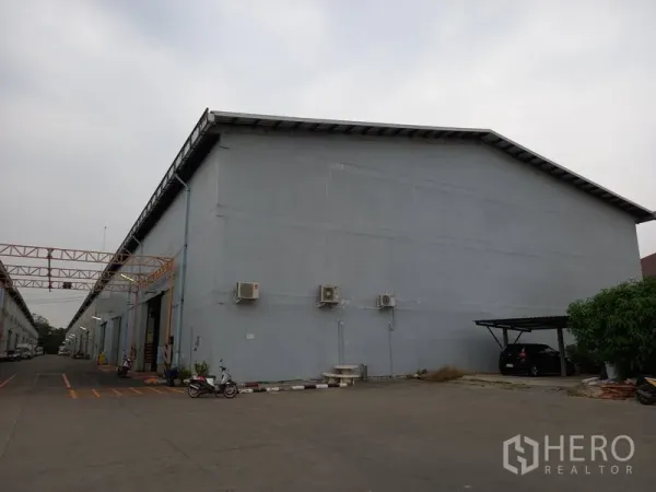Warehouse for rent in Mueang Pathum Thani, Pathum Thani - Exterior side of a high-wall warehouse with loading bay markings and a small carport within the compound.