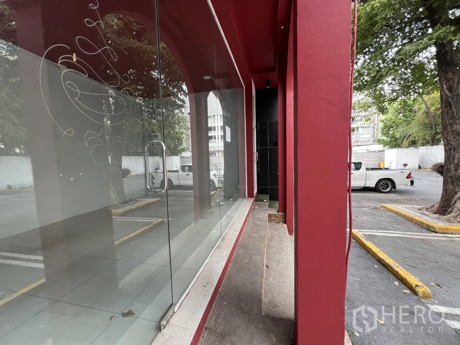 零售空间 for rent in Watthana, Bangkok - 近景玻璃门面与红色立柱，拉手清晰，可见停车区与绿树。
