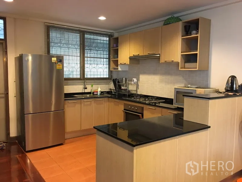 Detached House for rent in Khlong Toei, Bangkok - Modern U-shaped kitchen with black granite countertops, gas hob, oven and a stainless-steel refrigerator.