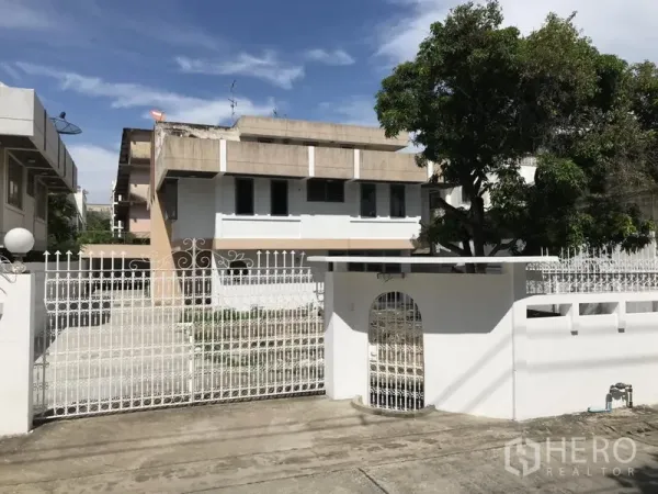 Detached House for rent or sale in Khlong Toei, Bangkok - Front gate view of a white two-storey house with metal fence, wide driveway and mature tree near BTS Phra Khanong.