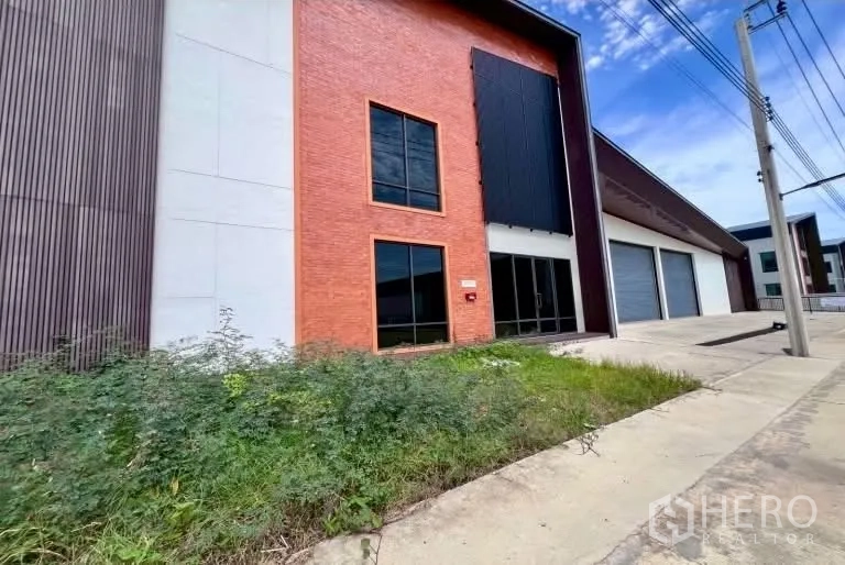 Warehouse for rent in Bang Bo, Samut Prakan - Front facade showing the office entrance, tall windows, and several roller shutters along the road.