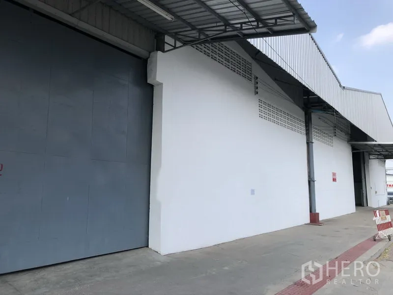 货栈 出租在Nonthaburi - 仓库外立面，设有大型灰色推拉门与带雨棚的装卸区，通道宽敞。