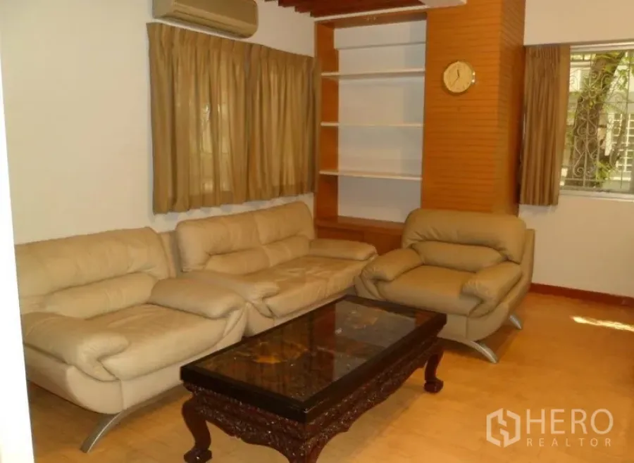 Condominium for rent in Pathum Wan, Bangkok - Bright living room with beige leather sofa set, carved coffee table, built-in shelving, and curtained window.