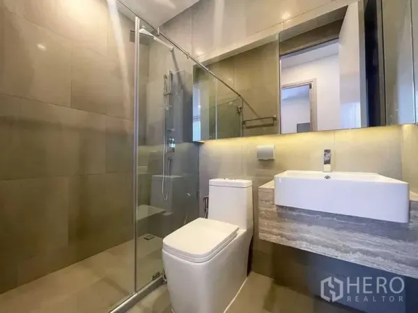 Condominium for rent in Ratchathewi, Bangkok - Modern bathroom featuring a glass-enclosed shower, wall-mounted sink, backlit mirror and tiled walls.