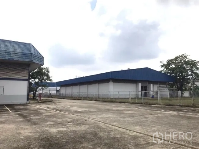 工厂 出租在Bang Bua Thong, Nonthaburi - 蓝顶仓库之间的开阔混凝土堆场与围栏。