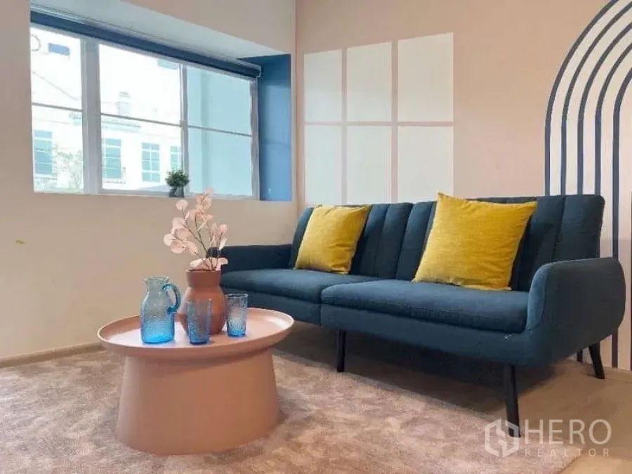 Townhouse for rent in Bang Bo, Samut Prakan - Cozy sitting area with blue sofa, yellow cushions and pastel coffee table by a bright window.