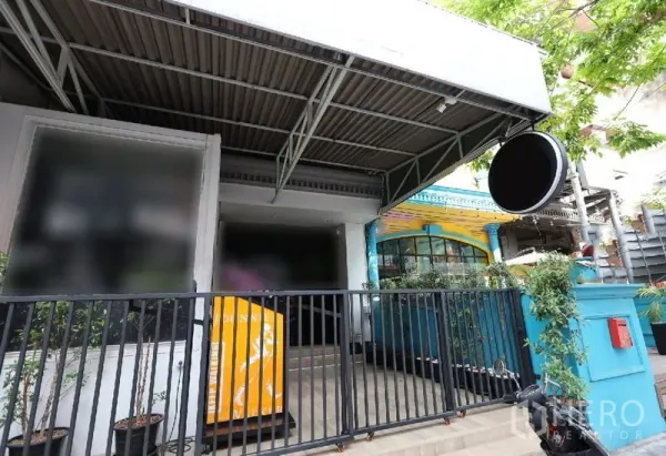 店屋 出租或出售在Watthana, Bangkok - 街铺外立面，金属雨棚、圆形招牌与黑色栅栏入口，周围绿植点缀
