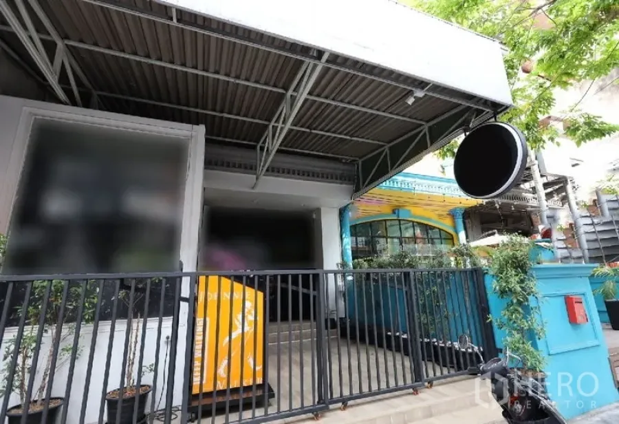 店屋 出租或出售在Watthana, Bangkok - 街铺外立面，金属雨棚、圆形招牌与黑色栅栏入口，周围绿植点缀