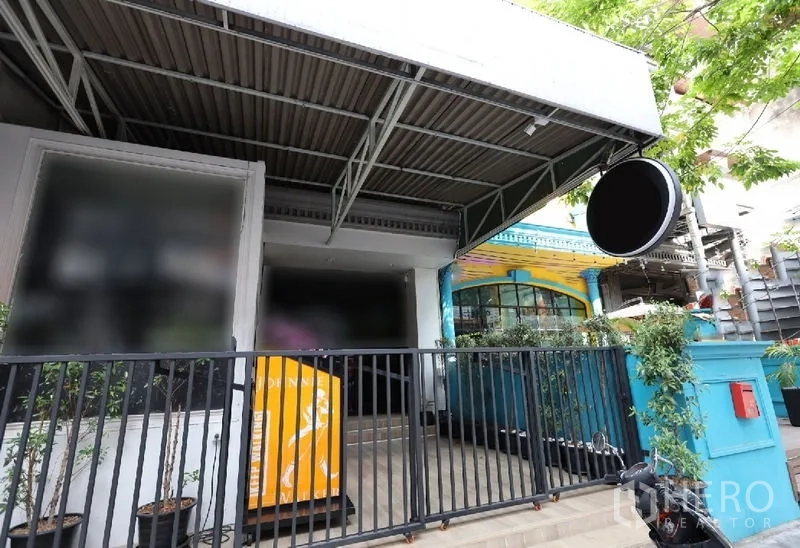 店屋 出租或出售在Watthana, Bangkok - 街铺外立面，金属雨棚、圆形招牌与黑色栅栏入口，周围绿植点缀