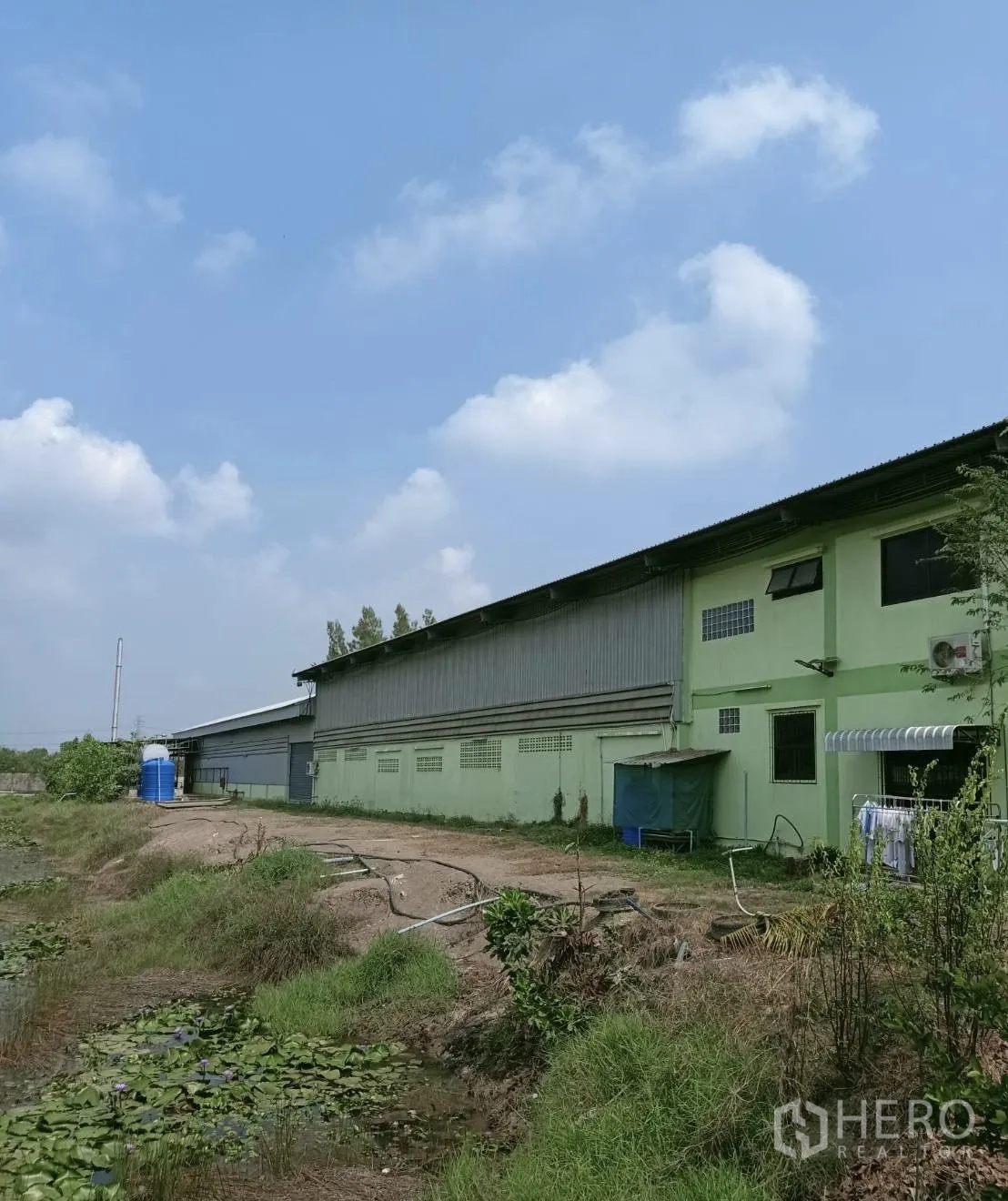 โรงงาน for rent or sale in Nong Suea, Pathum Thani - ด้านข้างอาคารโรงงานสีเขียวผนังเมทัลชีท มีทางบริการและสระน้ำจอกบัว พร้อมแทงก์น้ำสีน้ำเงินด้านท้ายอาคาร