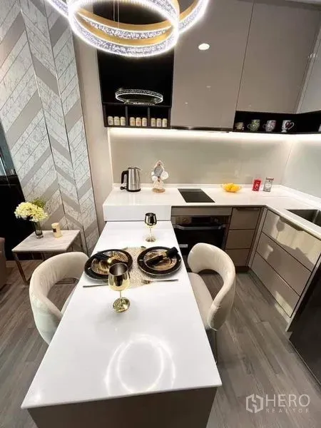 Condominium for sale in Bang Sue, Bangkok - Modern kitchenette with glossy cabinets, white L-shaped counter, two-seat dining bar and ring chandelier.