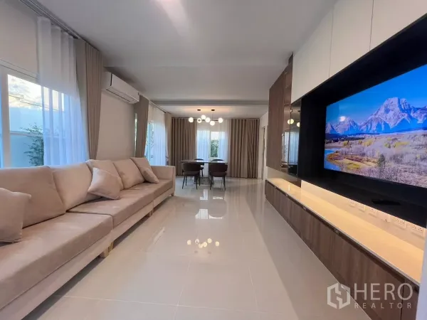 Detached House for rent in Bang Phli, Samut Prakan - Bright living room with long beige sofa, glossy floor tiles and wall-mounted TV facing the dining area.