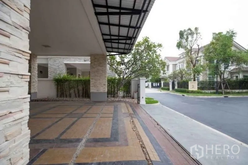 Detached House for rent or sale in Bang Phli, Samut Prakan - Covered carport and paved driveway looking onto a landscaped, gated community street.