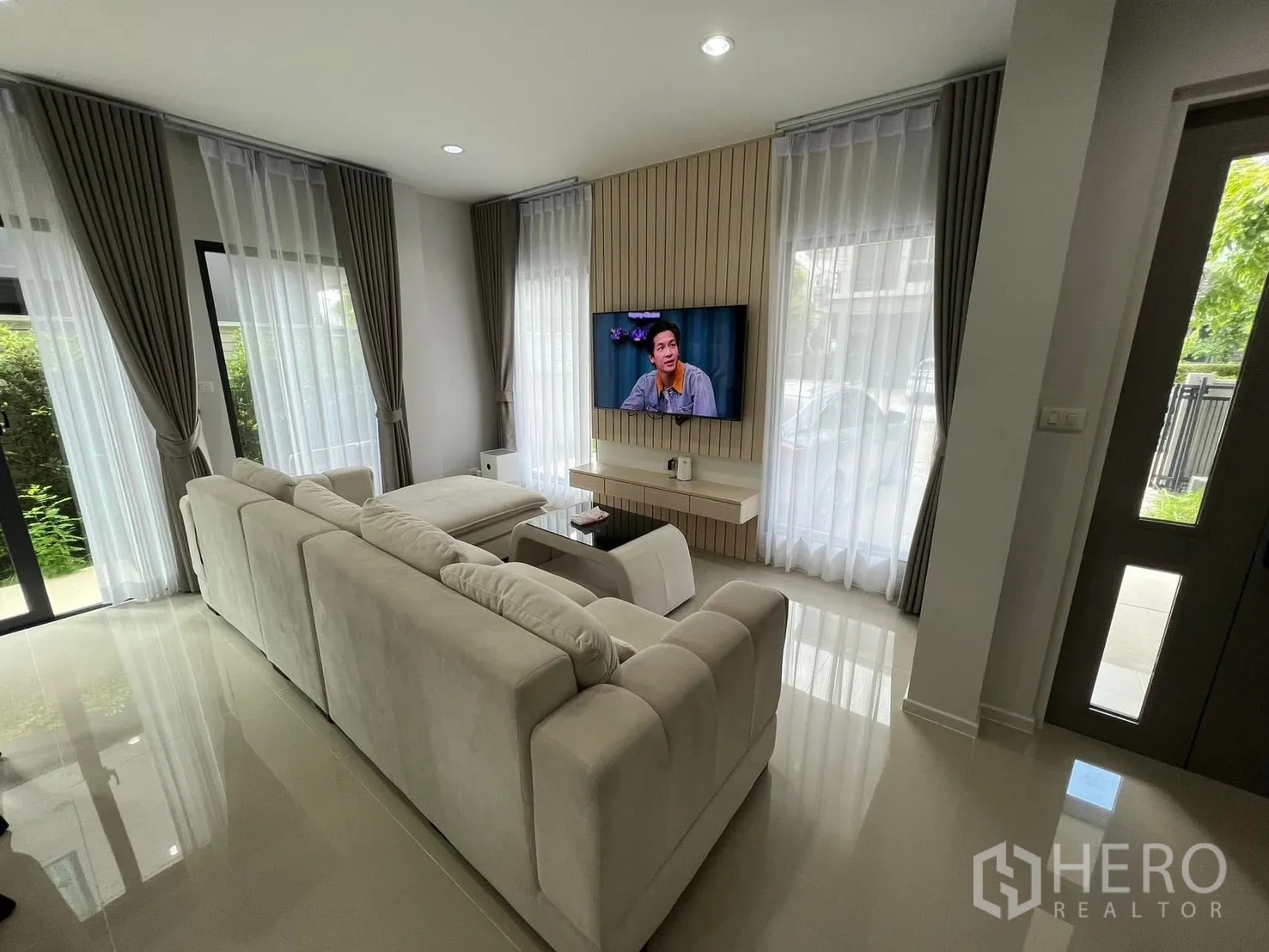 Semi-detached House for rent in Saphan Sung, Bangkok - Living room with sectional sofa, wall-mounted TV, and tall windows with sheer curtains.