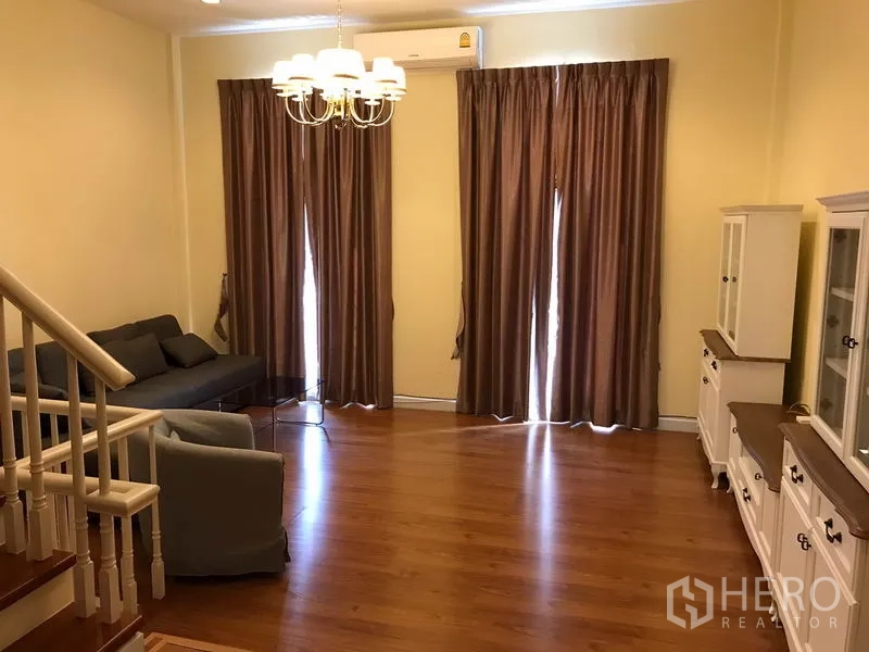 Townhouse for rent in Wang Thonglang, Bangkok - Spacious living room with wood floors, chandelier lighting, brown drapes, white display cabinets and a staircase.