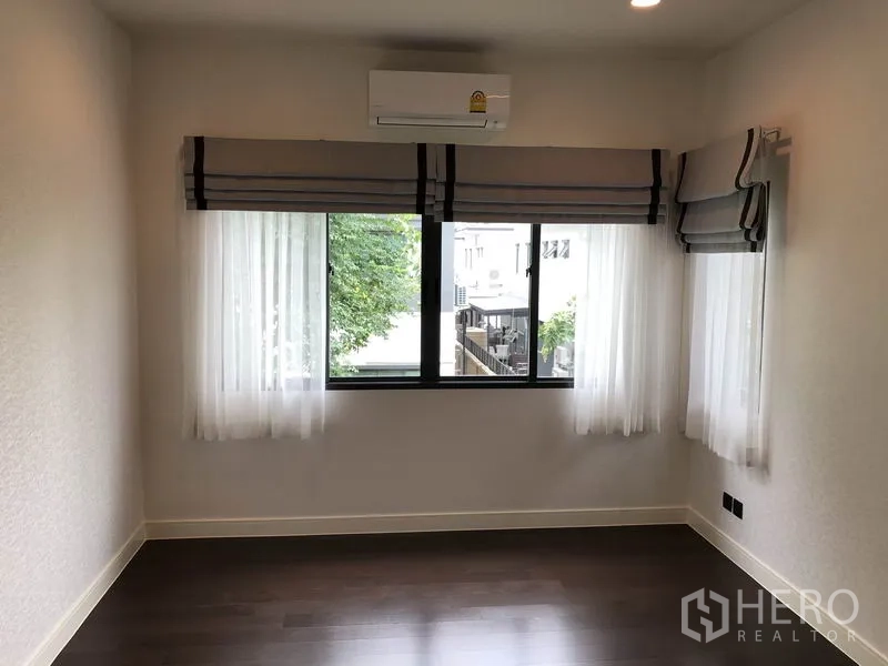 Detached House for rent or sale in Bang Kapi, Bangkok - Corner bedroom with large windows, roman blinds, sheer curtains and wall-mounted air conditioner.