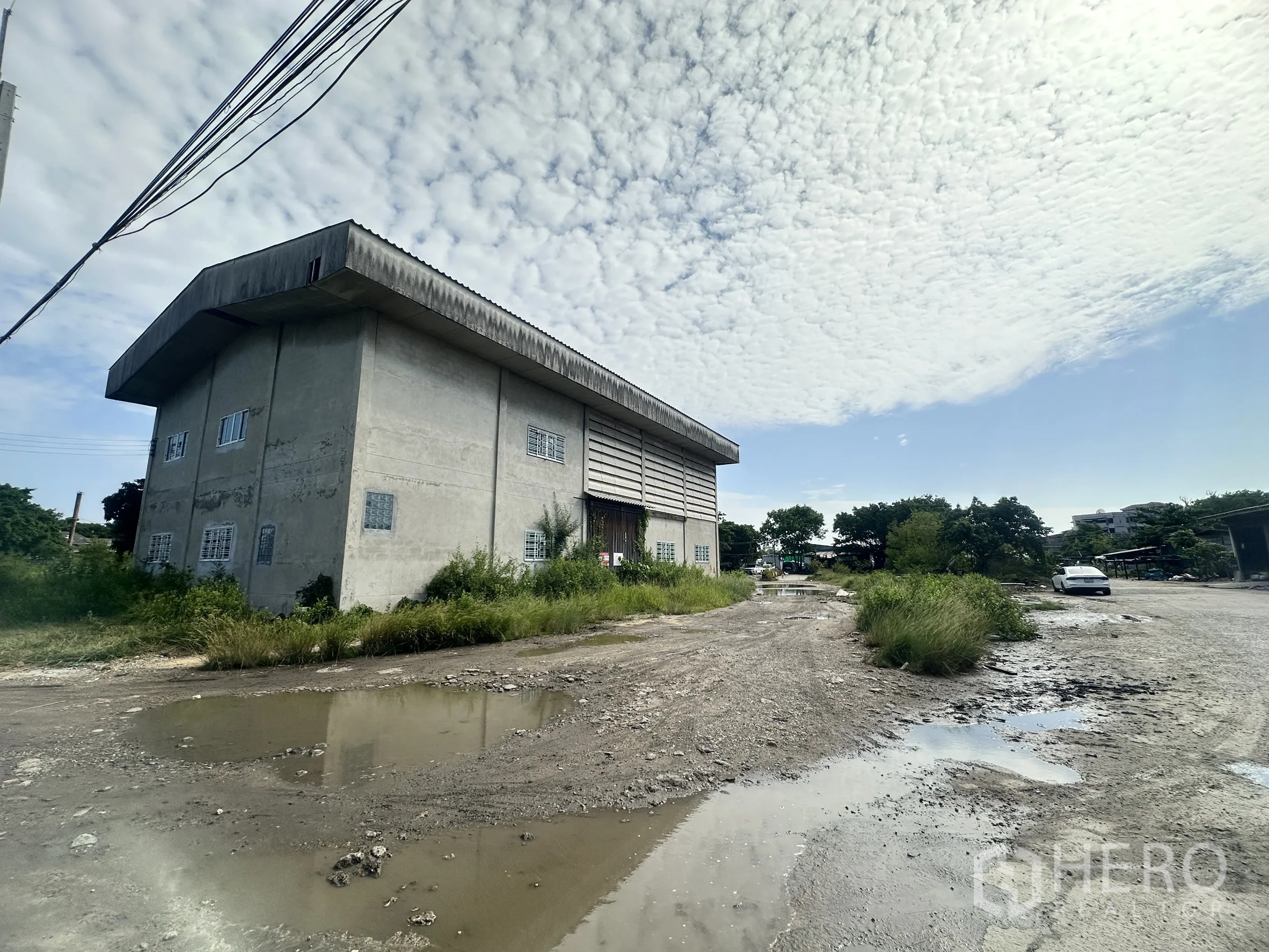 Land for sale in Mueang Samut Prakan, Samut Prakan - Exterior view of the warehouse beside an unpaved access road with surrounding vegetation.