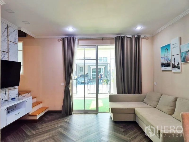 联排别墅 出租或出售在Phra Pradaeng, Samut Prakan - 客厅配L型沙发、电视墙，推拉玻璃门通向前庭。