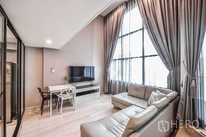 Condominium for rent in Sathon, Bangkok - Bright living room with L-shaped leather sofa, TV console and dining table under floor-to-ceiling windows.