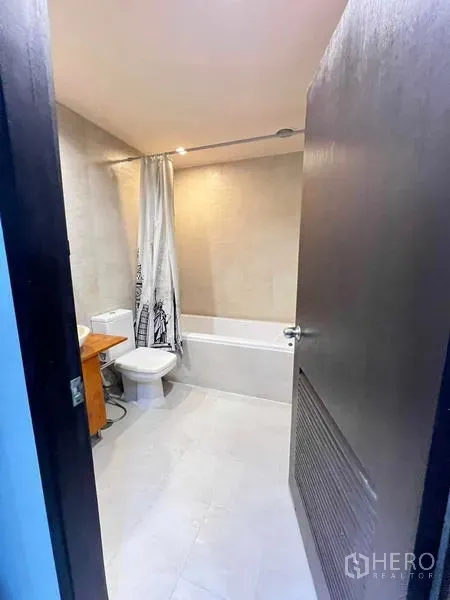 Condominium for rent in Ratchathewi, Bangkok - Bathroom view from doorway showing the tub with curtain and a wooden vanity.