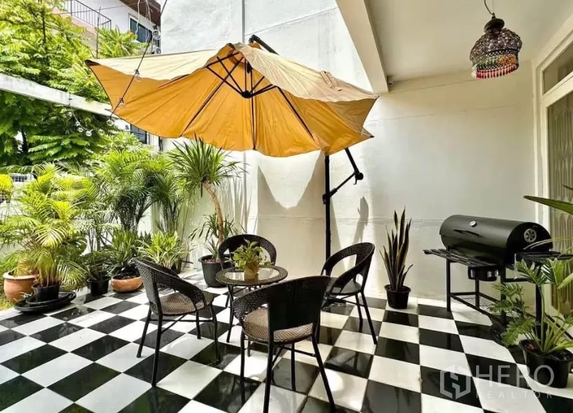 Townhouse for rent in Watthana, Bangkok - Covered terrace with table, chairs, large umbrella, potted plants and barbecue grill on checker tiles.