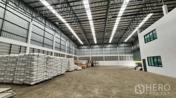 货栈 出租在Ban Phaeo, Samut Sakhon - 明亮的仓库通道，屋顶采光板充足，墙边堆放托盘袋装货物。