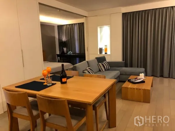 Condominium for rent in Watthana, Bangkok - Open-plan living and dining area with wooden table, L-shaped sofa and large drapes.