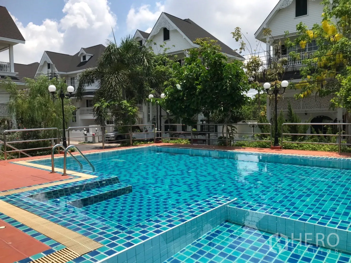 Detached House for rent in Mueang Samut Prakan, Samut Prakan - Outdoor communal swimming pool with tiled deck surrounded by greenery and houses.