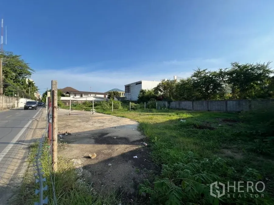 ที่ดิน ขายในBang Na, Bangkok - มุมริมถนนแสดงทางเข้าพื้นเทคอนกรีต หลักเขต และอาคารกับต้นไม้โดยรอบ