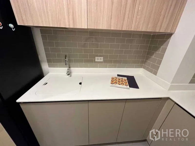 公寓 出租在Ratchathewi, Bangkok - 紧凑厨房，白色台面、带盖水槽、浅木柜与灰色地铁砖背景墙。